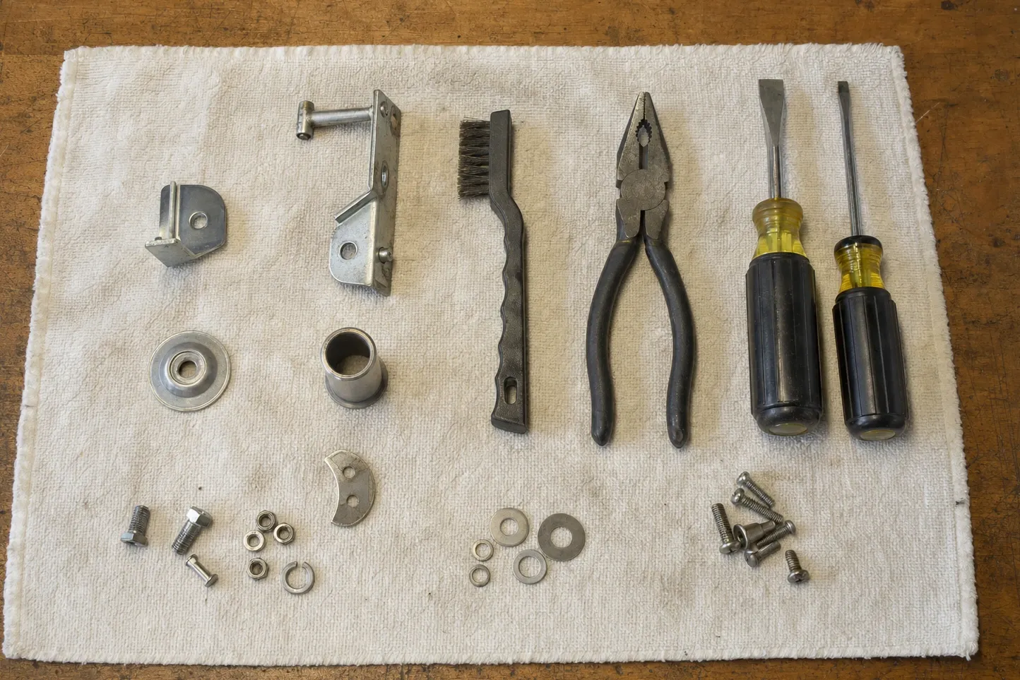 Hinge brackets, a bushing, a bearing race, bolts and washers set out in rows on a cloth beside pliers and two screwdrivers