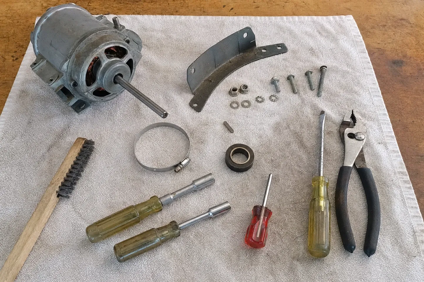 Open-frame electric motor on a cloth with its rotor visible through the end bell and a long plain shaft, beside a curved mounting strap, a rubber-lined bushing, bolts, a wire brush, nut drivers and pliers. Numbered marks are drawn over it, at the rotor visible through the end bell, rubber-lined bushing and the curved mounting strap.