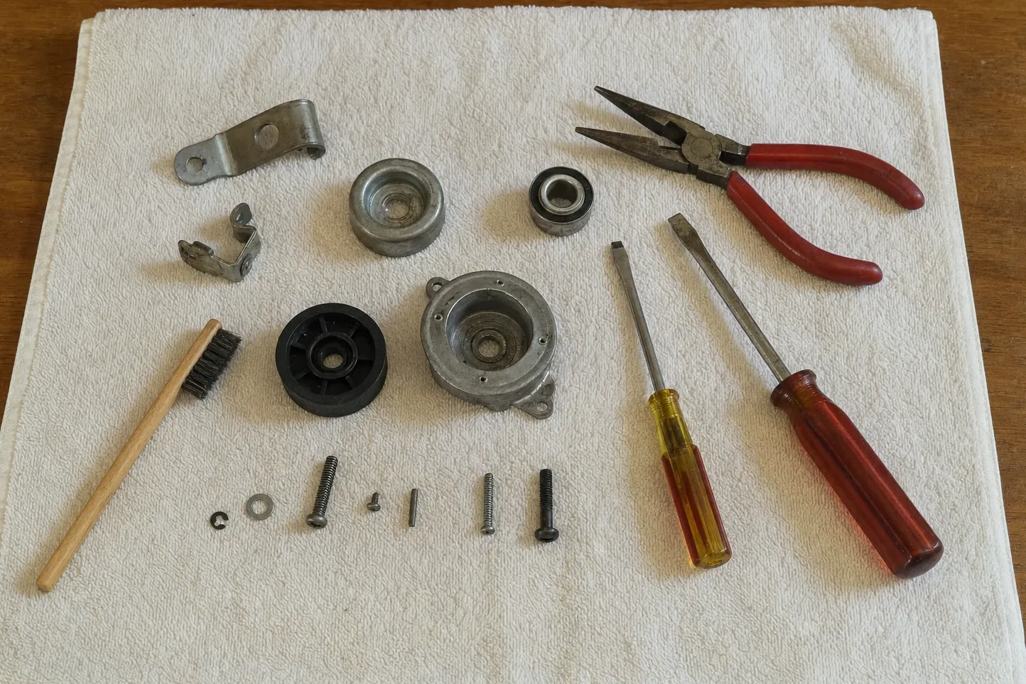 Sealed ball bearing, a cast bearing housing with its cover off, a black nylon wheel and an alloy roller set out on a towel with a spring clip, screws, a wire brush, screwdrivers and needle-nose pliers. Numbered marks are drawn over it, at the bearing housing with the cover off, sealed ball bearing and the plain alloy roller.